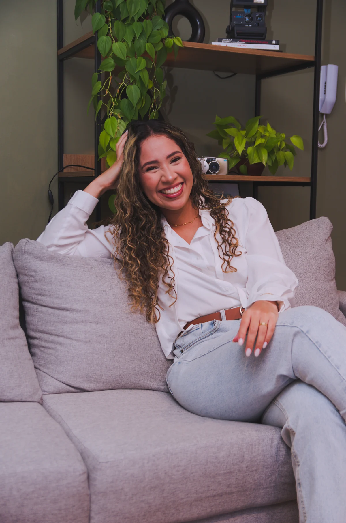 Poli Fernandes sorrindo, sentada no sofá de camisa branca, com plantas ao fundo.