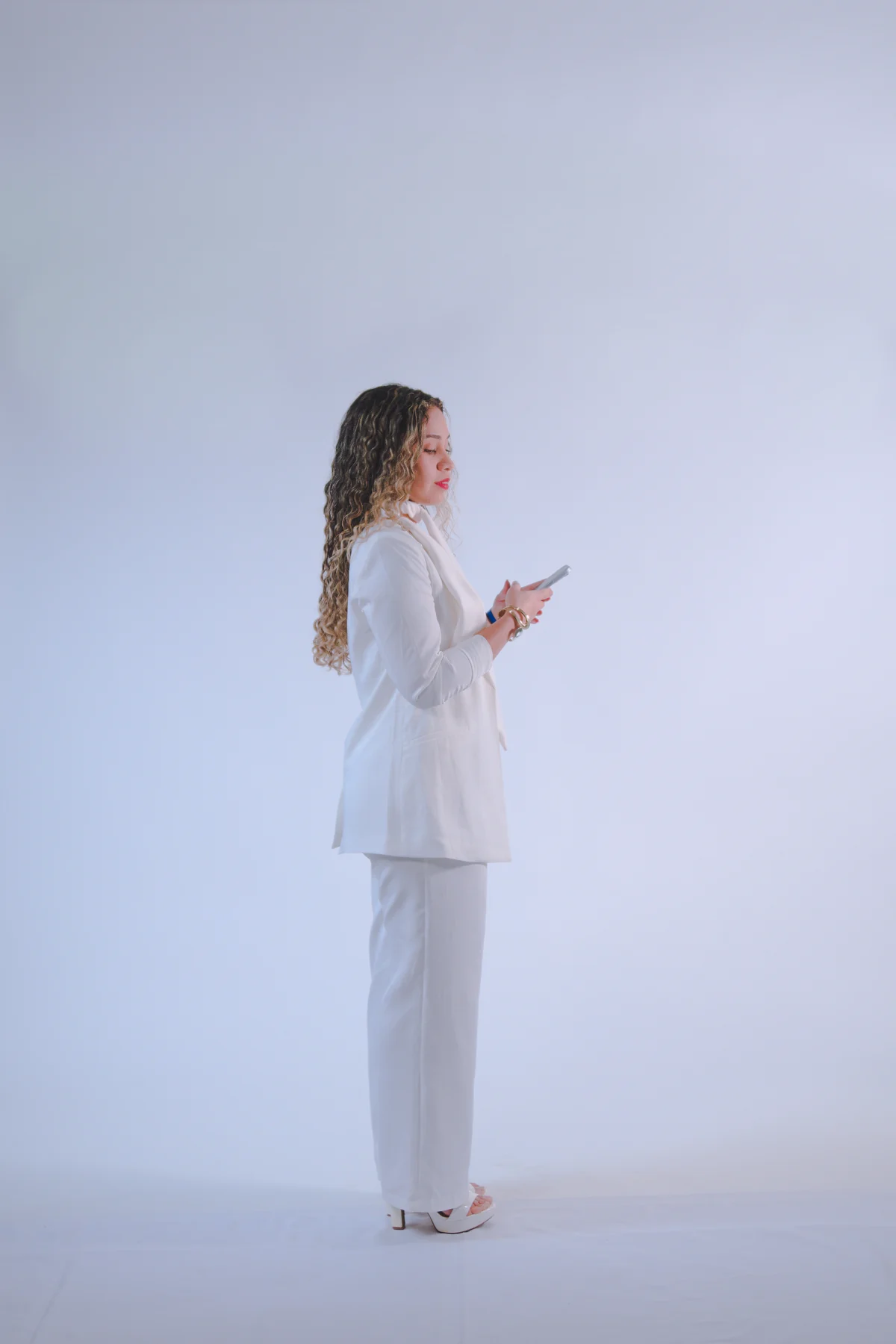 Poli Fernandes de branco, em pé e de perfil, em um estúdio de fundo claro.